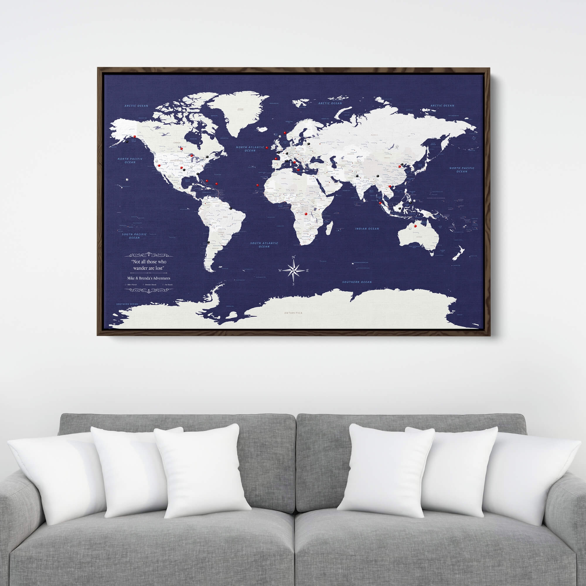 Navy Explorers world travel map canvas with red push pins in dark walnut frame above gray sofa